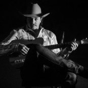 Black & white image of a man sat holding a guitar wearing a cowboy hat, shirt, jeans and boots